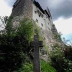 An image of Bran Castle perched on a cliff with a cross standing tall in front of it.